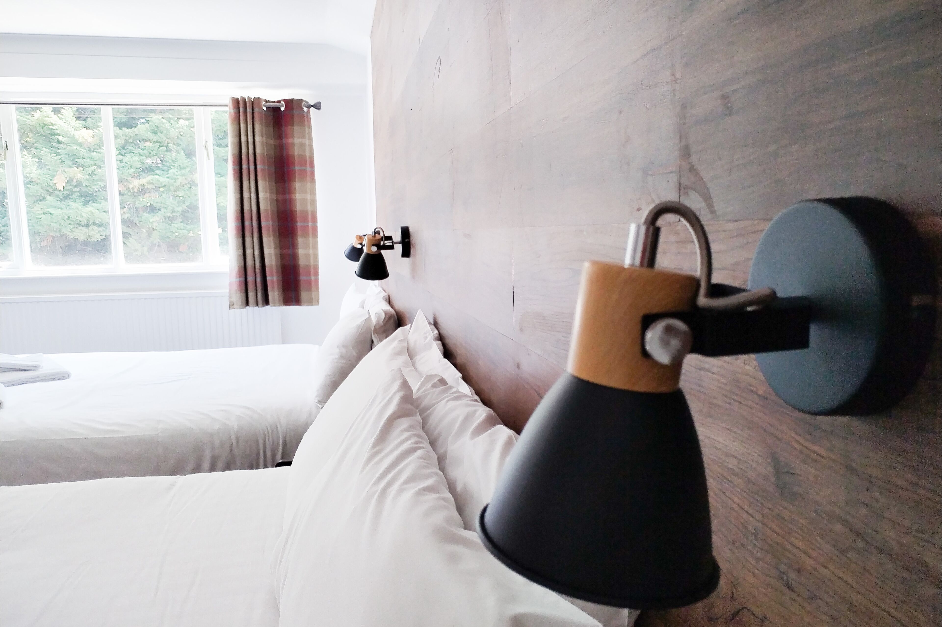 A bedroom with a wooden headboard, a bedside lamp, a window with curtains, and a bed with white bedding.