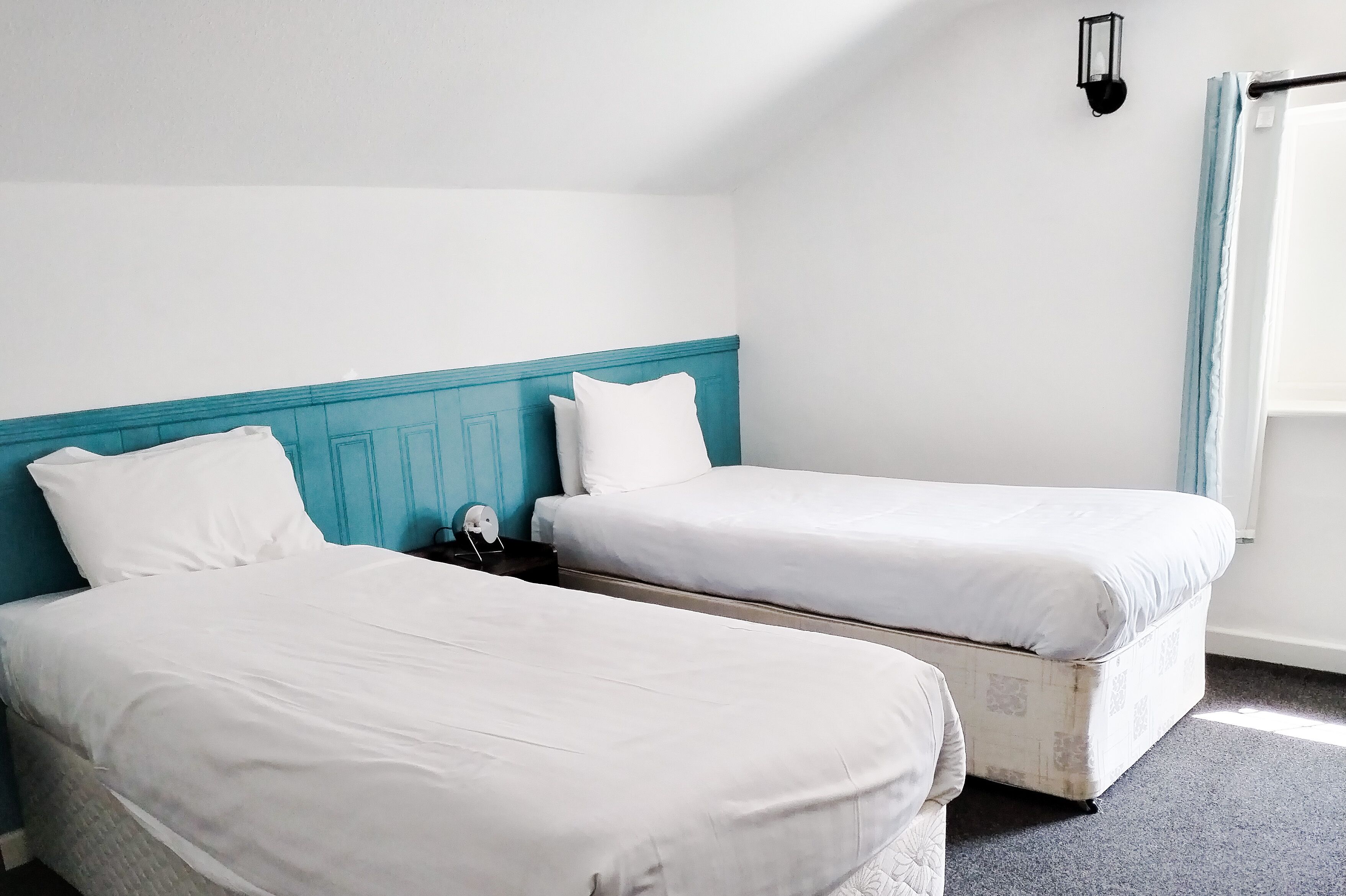 A room with two single beds, a teal headboard, white bedding, a nightstand with a lamp, and a window with a curtain.