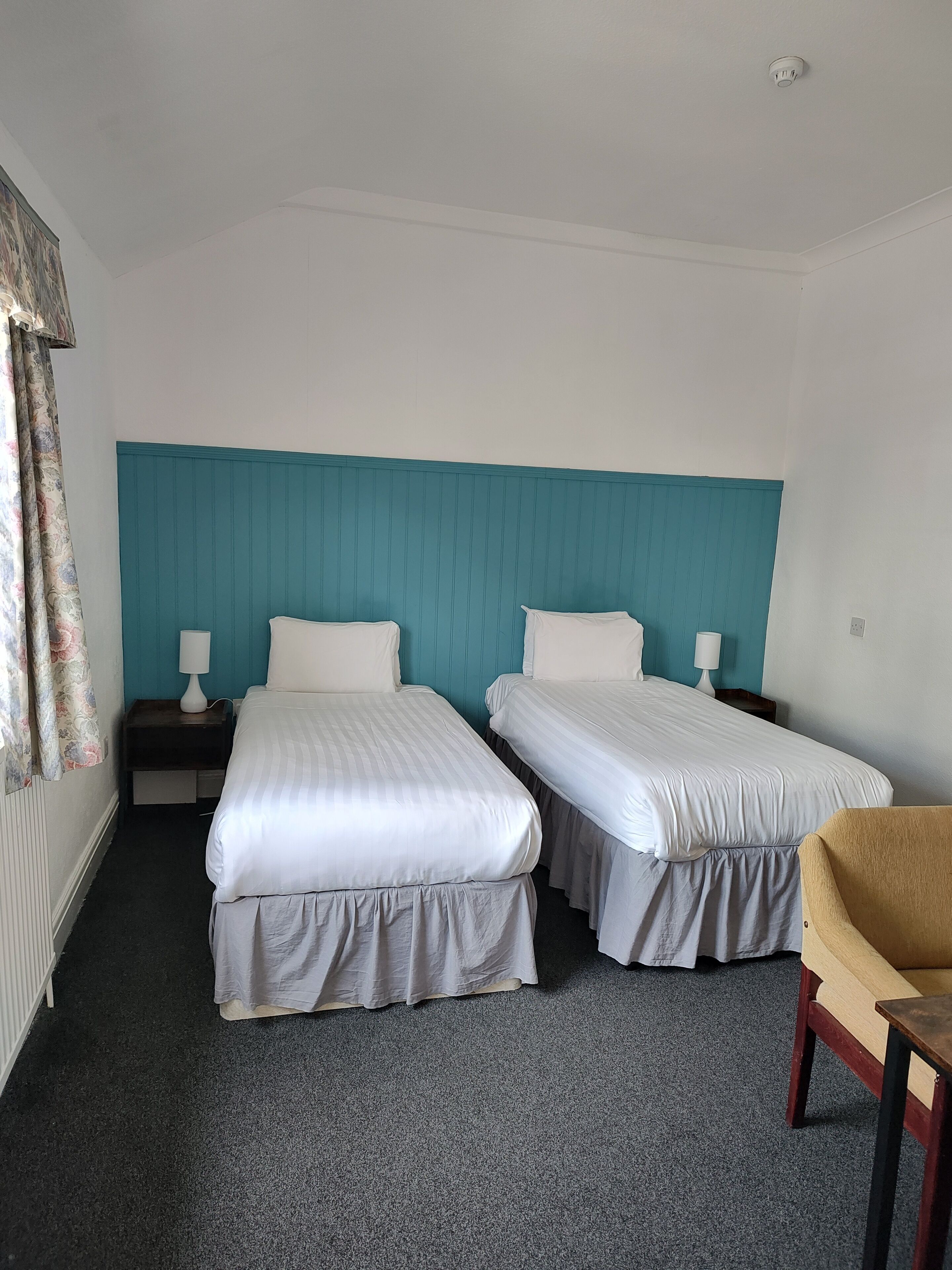 A hotel room with two single beds, a teal accent wall, a small wooden chair, and a nightstand with a lamp.