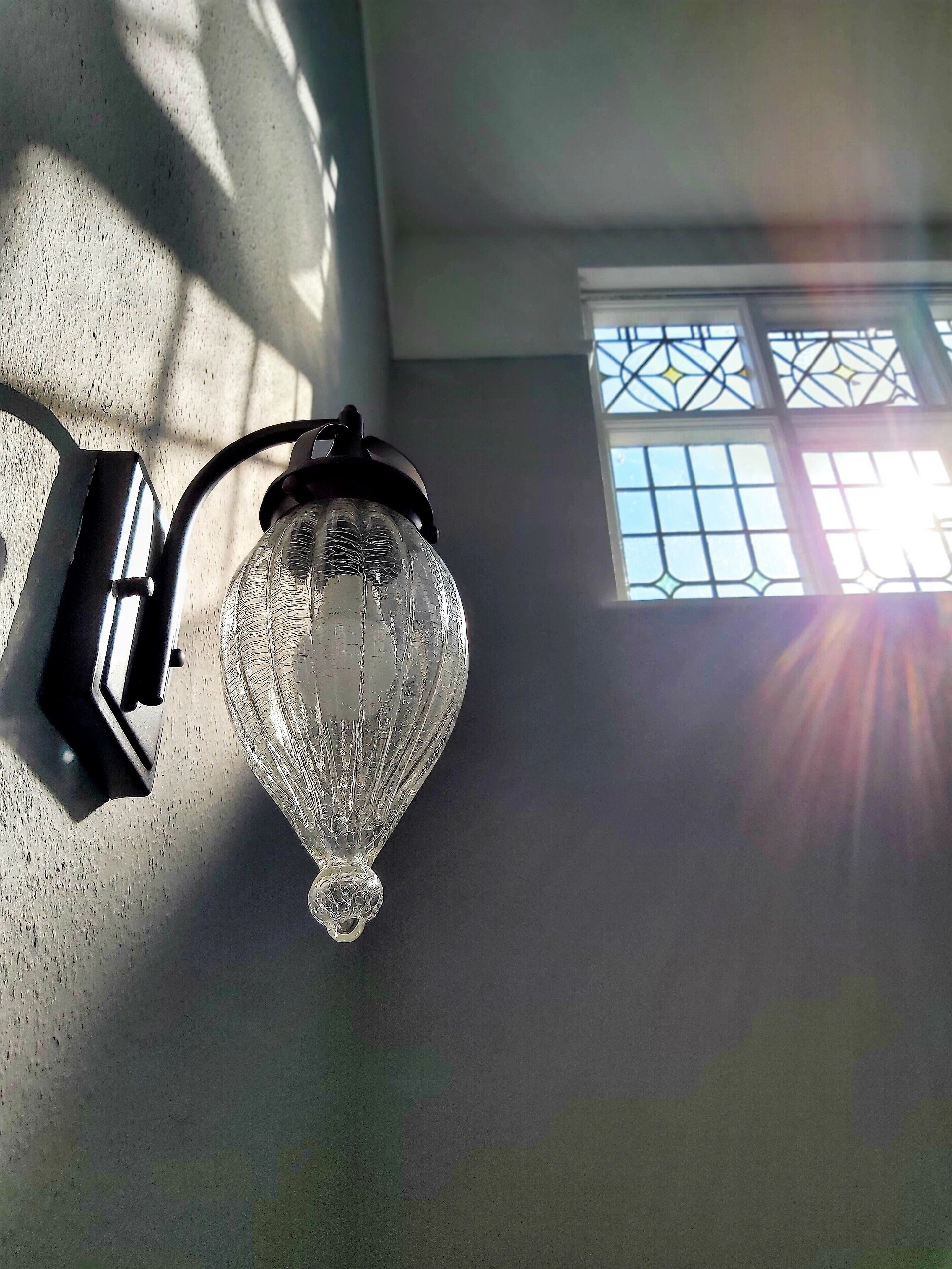A wall-mounted light fixture with a clear glass design, positioned next to a window with a grid pattern.