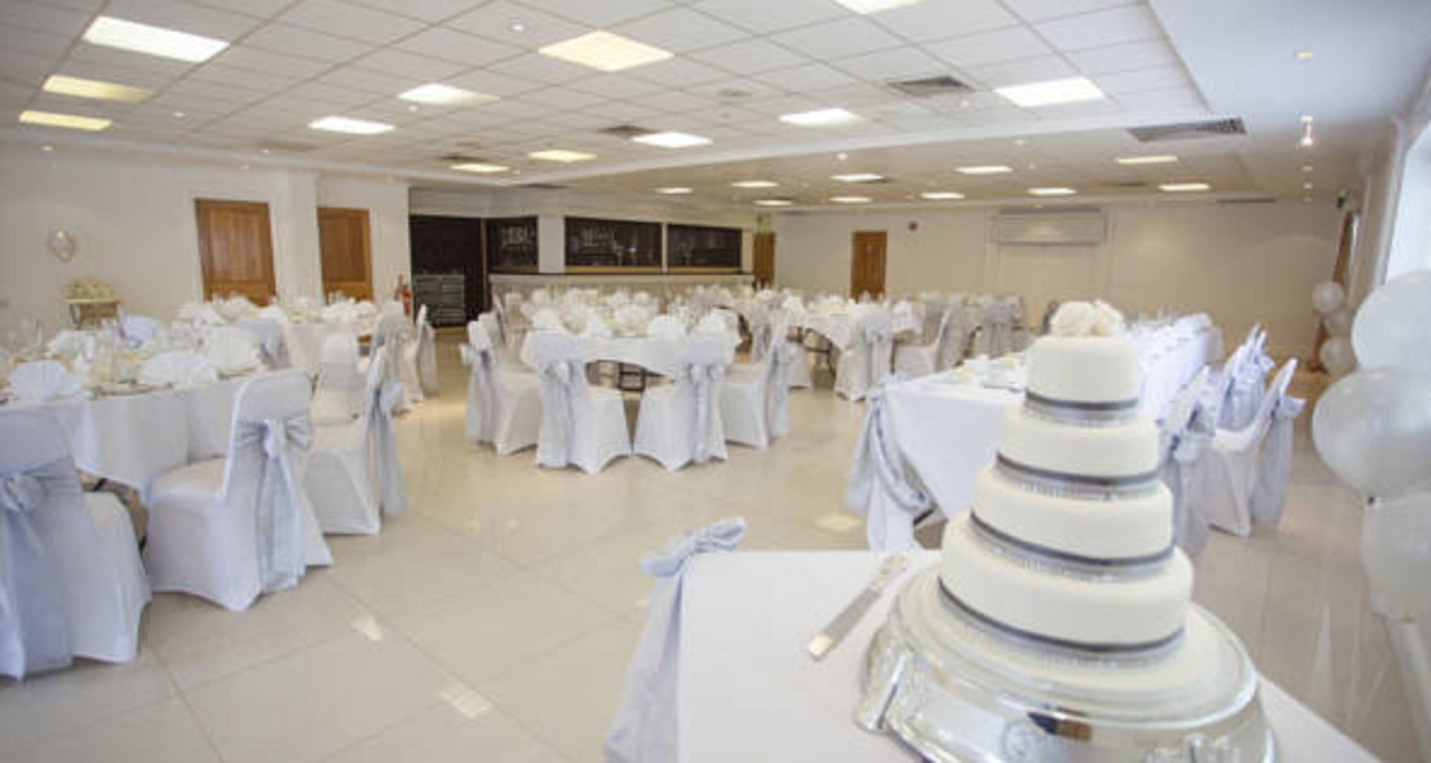 A banquet hall with round tables covered in white tablecloths and chairs, a tiered wedding cake, and a large chalkboard on the wall.