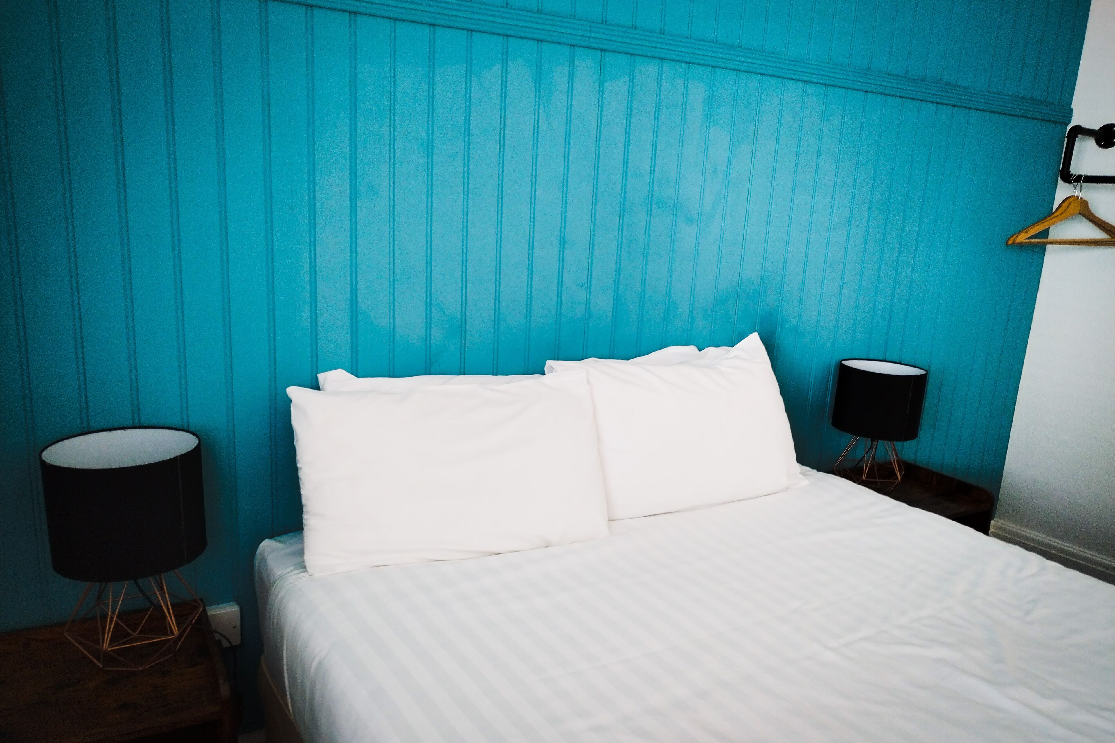 Ein Hotelzimmer mit Bett, weißer Bettwäsche, einer schwarzen Lampe auf einem Holznachttisch und einer wandfarbenen Wand in Türkis.
