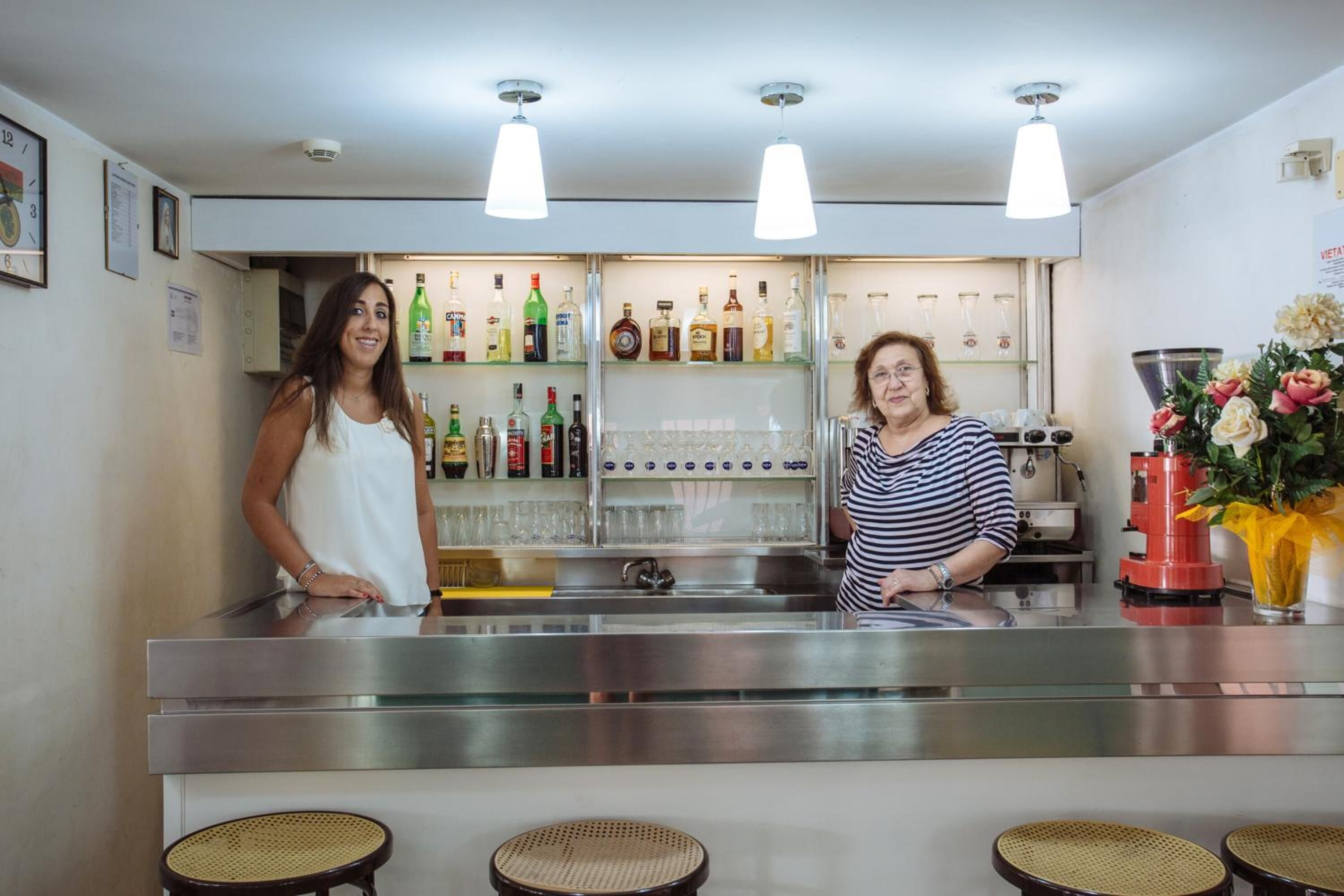 A bar area with a stainless steel counter, shelves with bottles, glasses, and a red machine. There are wicker stools in front.