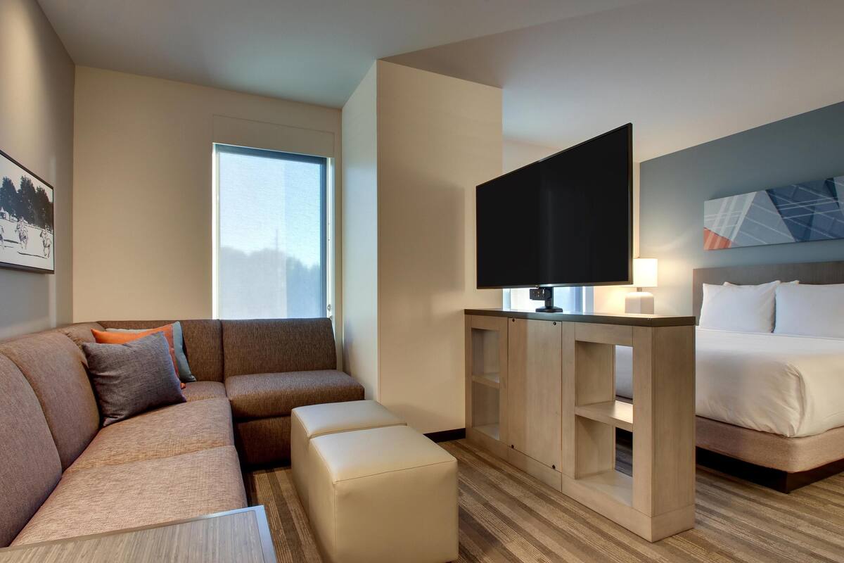 A hotel room with a sofa, ottoman, TV, bedside table, and a bed.