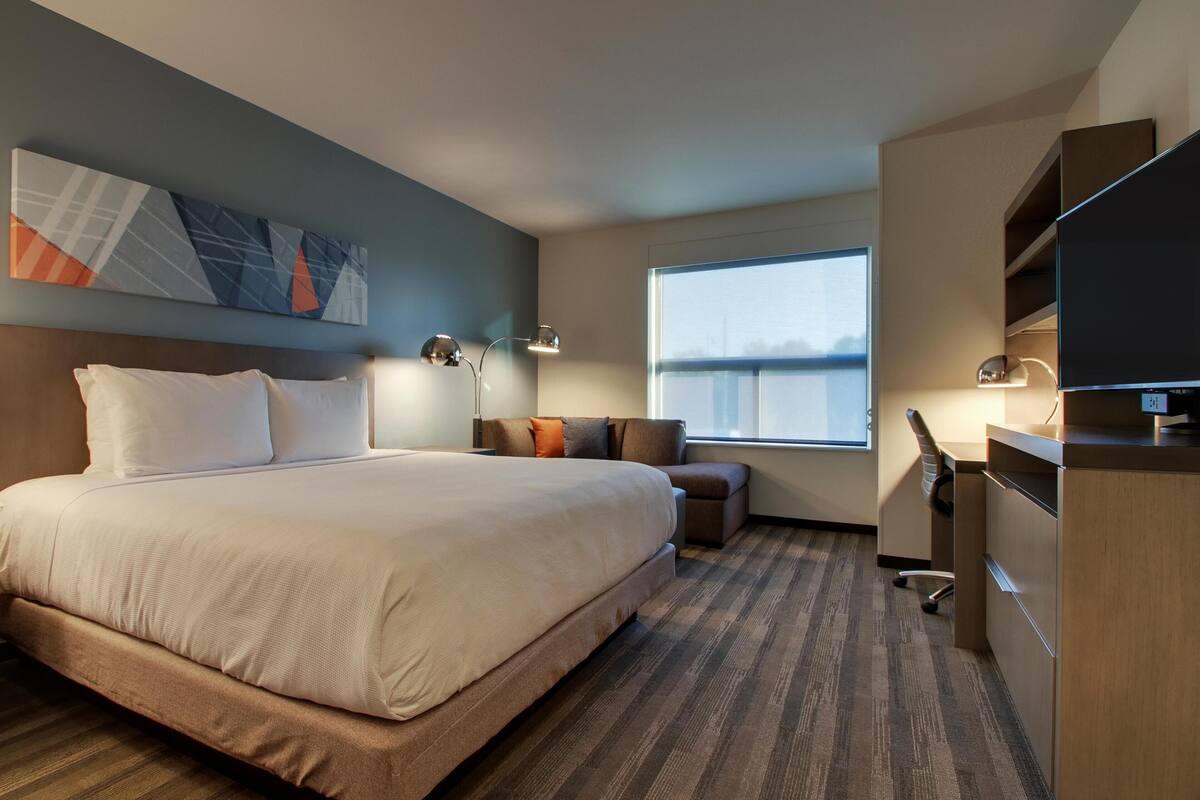 A hotel room with a large bed, a desk, a chair, a sofa, a TV, and a window with a view.