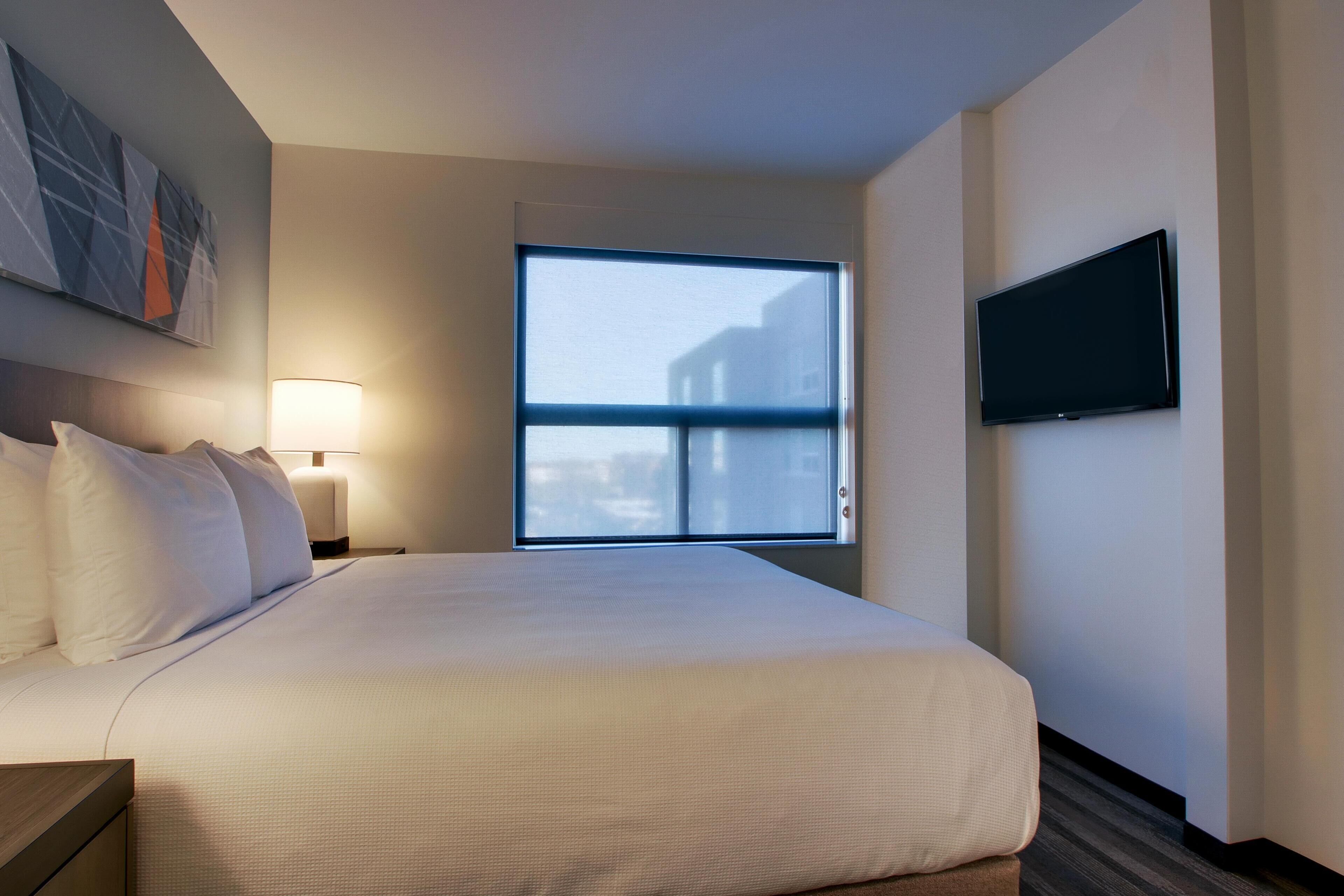 Ein Hotelzimmer mit Bett, Wandfernseher, Fenster und Nachttisch mit Lampe.