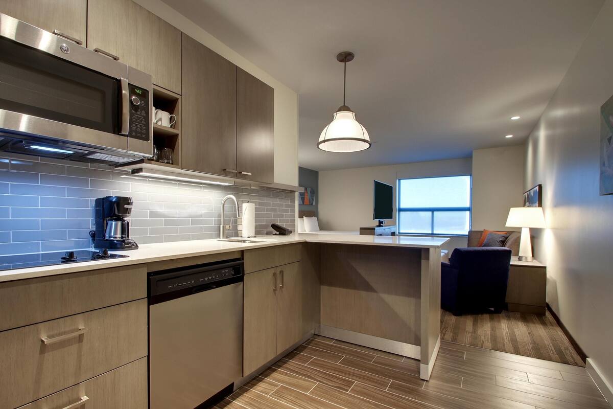 A modern kitchen with a built-in microwave, coffee maker, and dishwasher. There is a dining area with a chair and a table, and a window providing a view.