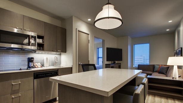 A modern kitchen with a dishwasher, microwave, and built-in oven. There is a dining area with a large island and bar stools. A flat-screen TV is mounted on the wall.