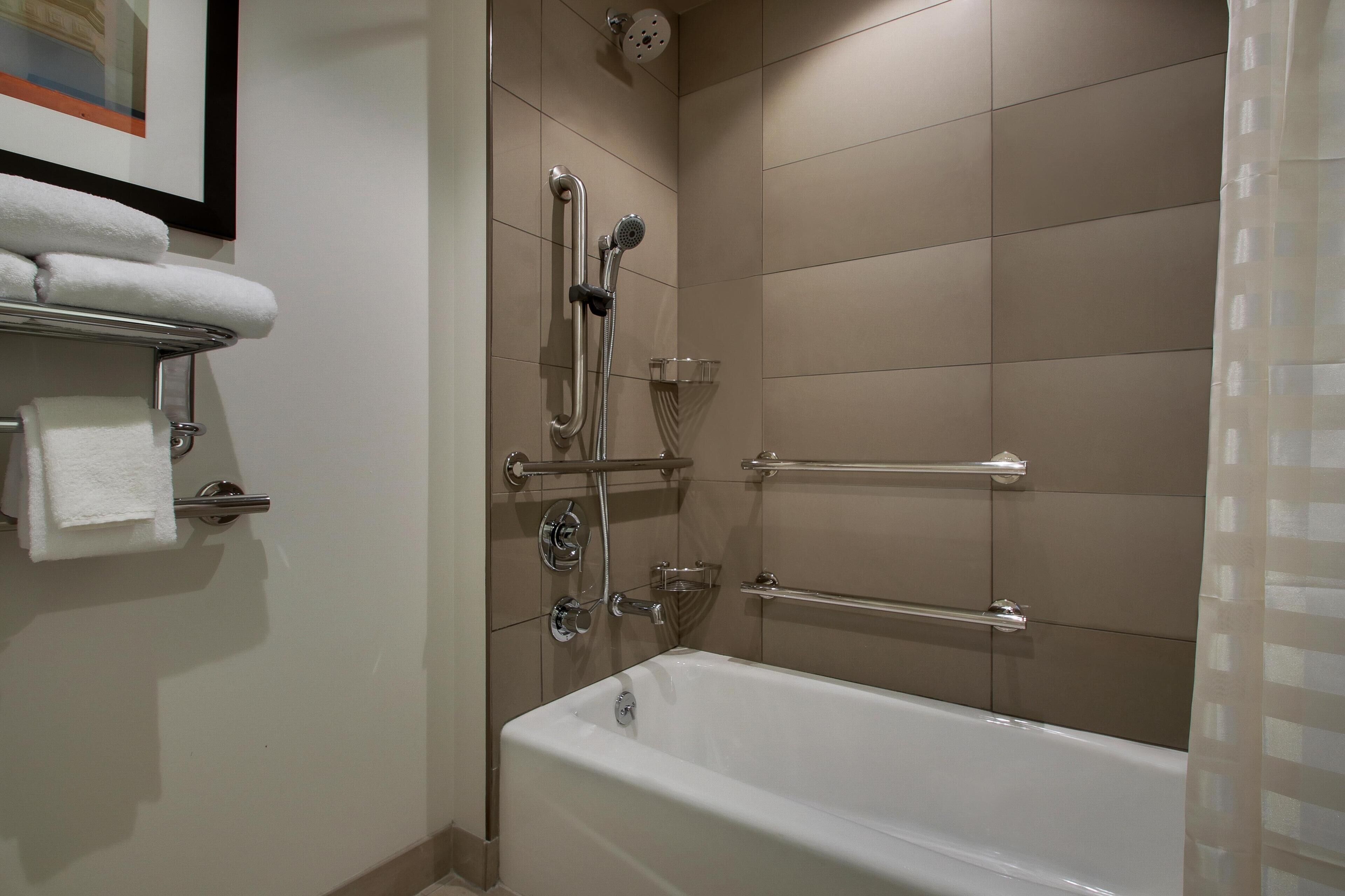 Ein Badezimmer mit Badewanne, Dusche inklusive Haltegriffen und einem Handtuchhalter mit gefalteten Handtüchern.