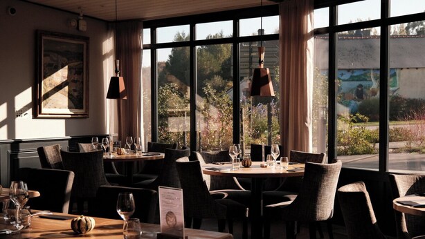 Um restaurante com mesas postas, amplas janelas com vista para o verde e iluminação pendente.