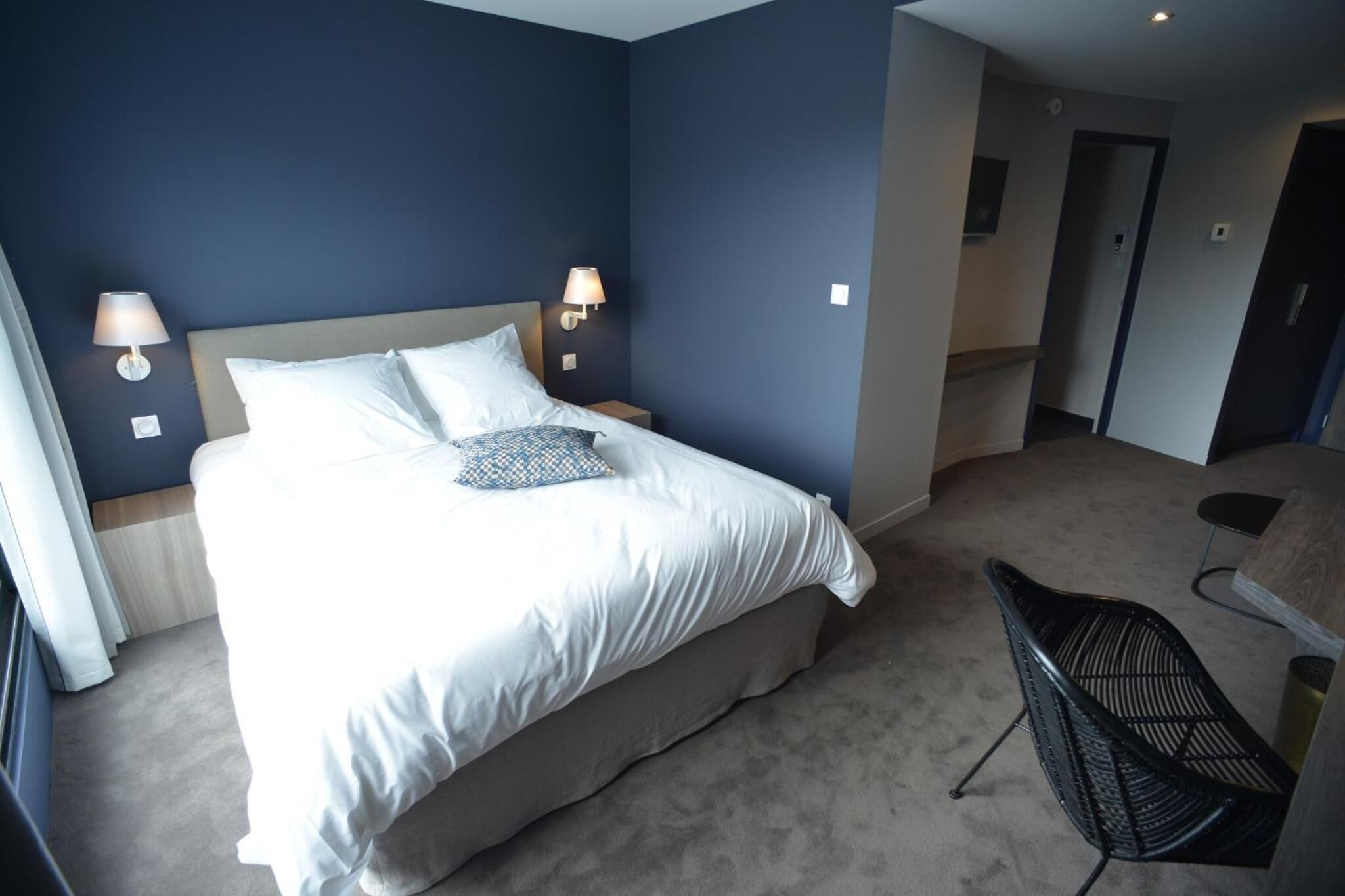 A hotel room with a large bed, two bedside lamps, a chair, and a door leading to another room.