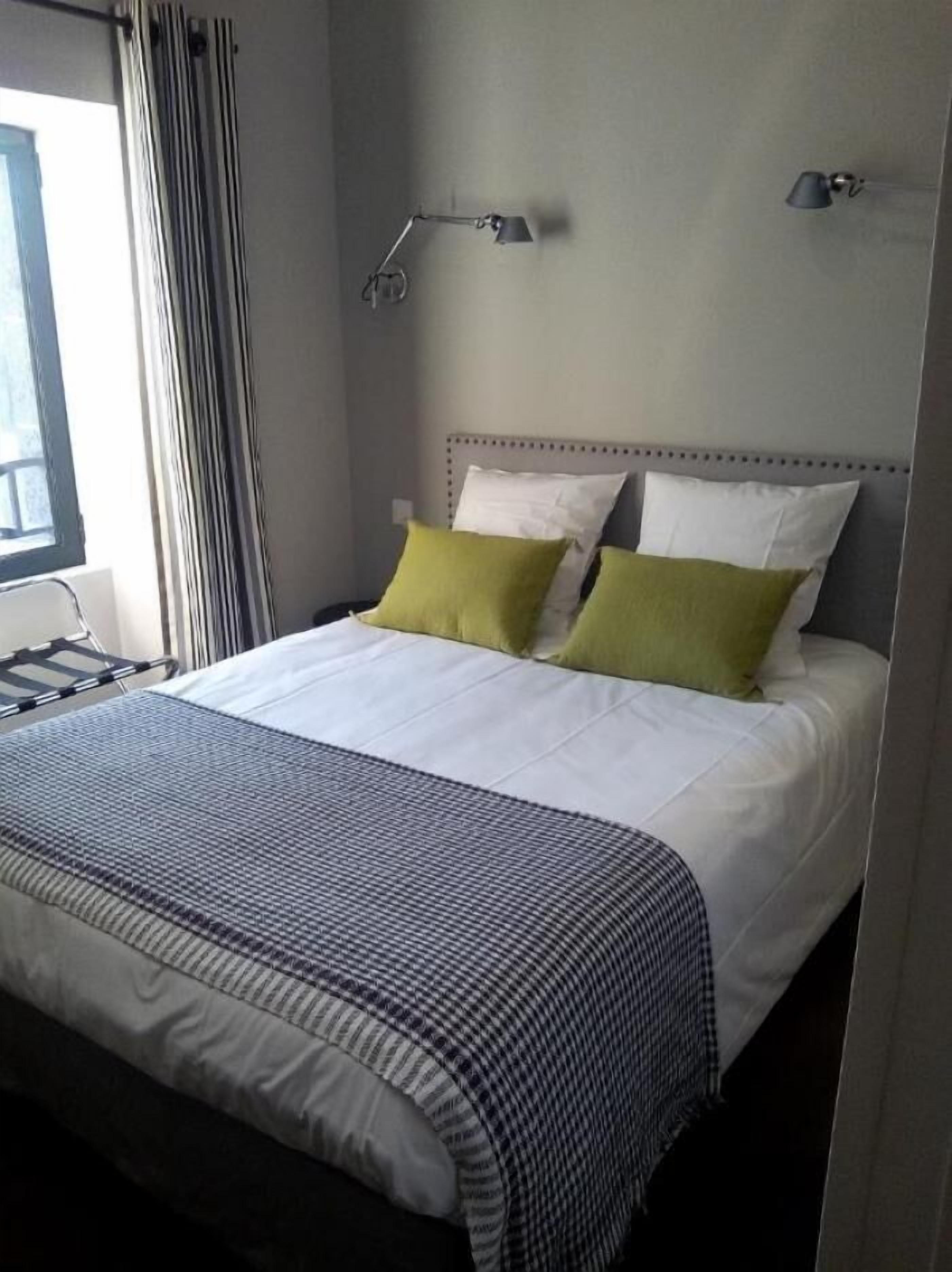 A neatly made bed with white and green pillows, a grey headboard, and a grey and white striped blanket. There are two bedside lamps on the wall.