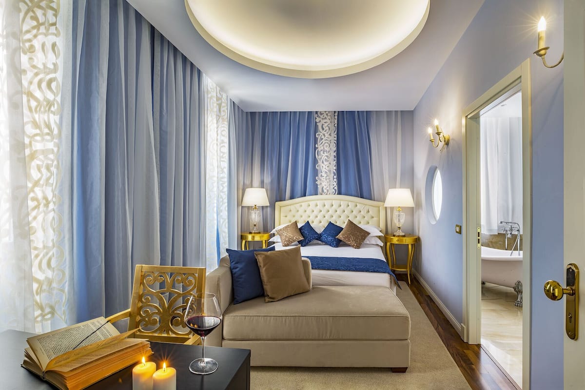 Een slaapkamer met een groot bed, twee nachtkastjes met lampen, een zithoek met een boek en kaarsen, en een badkamer die door een open deur zichtbaar is.