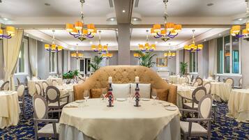A formal dining room with round tables, chairs, chandeliers, and a large upholstered sofa.