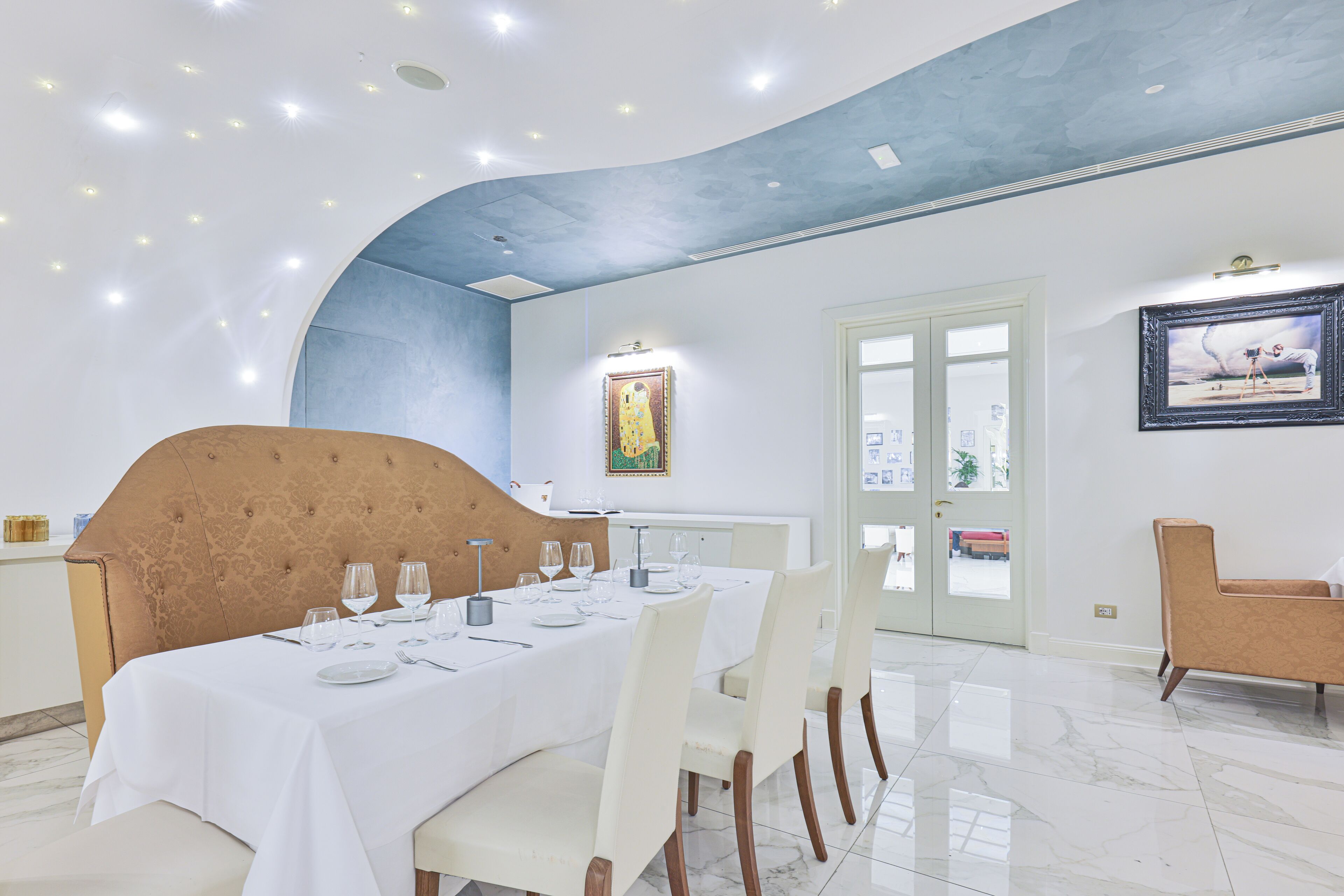 Zona pranzo con un lungo tavolo apparecchiato per un pasto, soffitto alto con illuminazione a incasso e porta con pannelli di vetro.