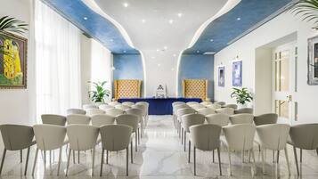 Sala de conferência com fileiras de cadeiras voltadas para um púlpito, paredes brancas e detalhes no teto em azul.