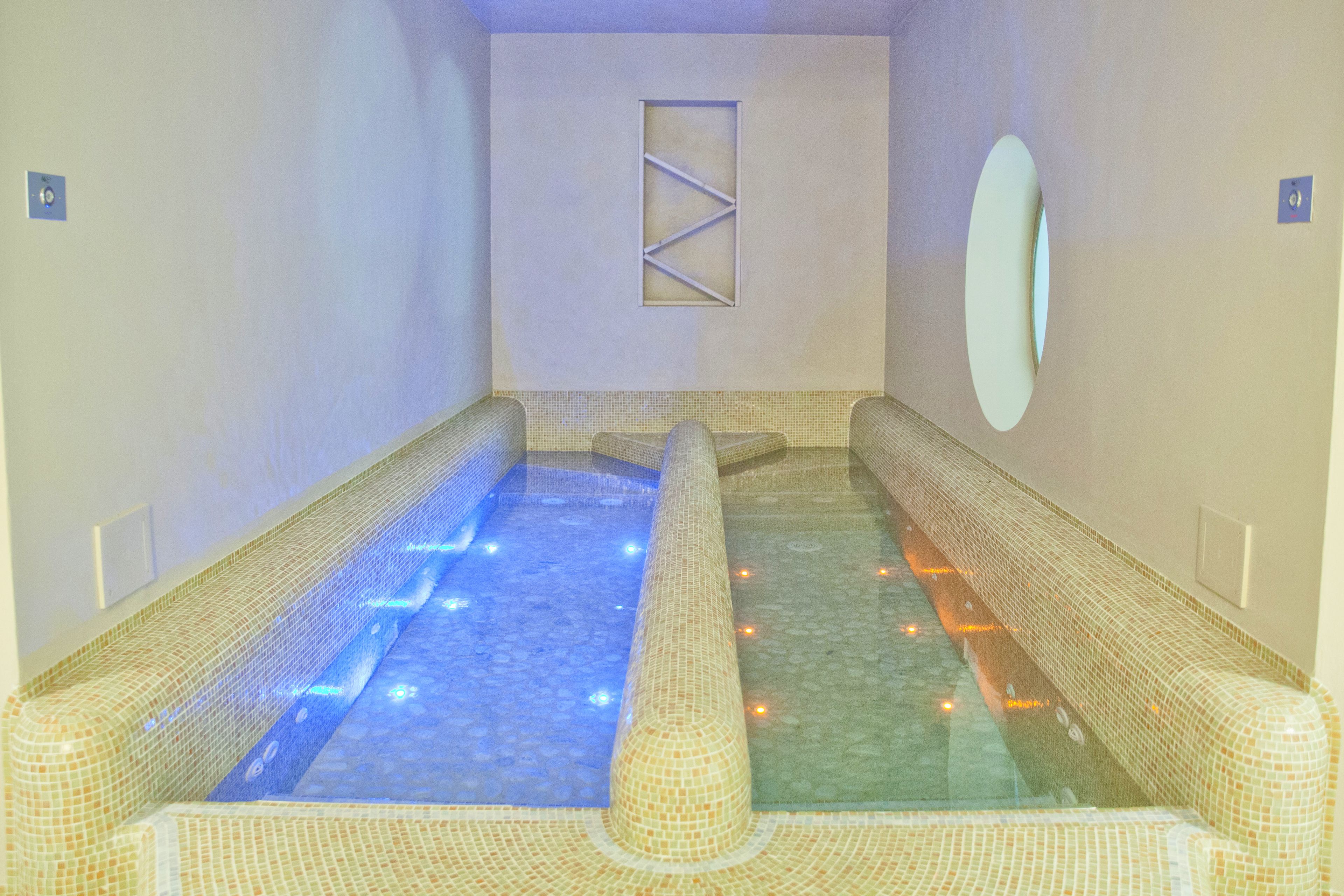 Une salle de spa dotée d’un jacuzzi entouré de parois en verre, de murs recouverts de mosaïques et d’une petite fenêtre.
