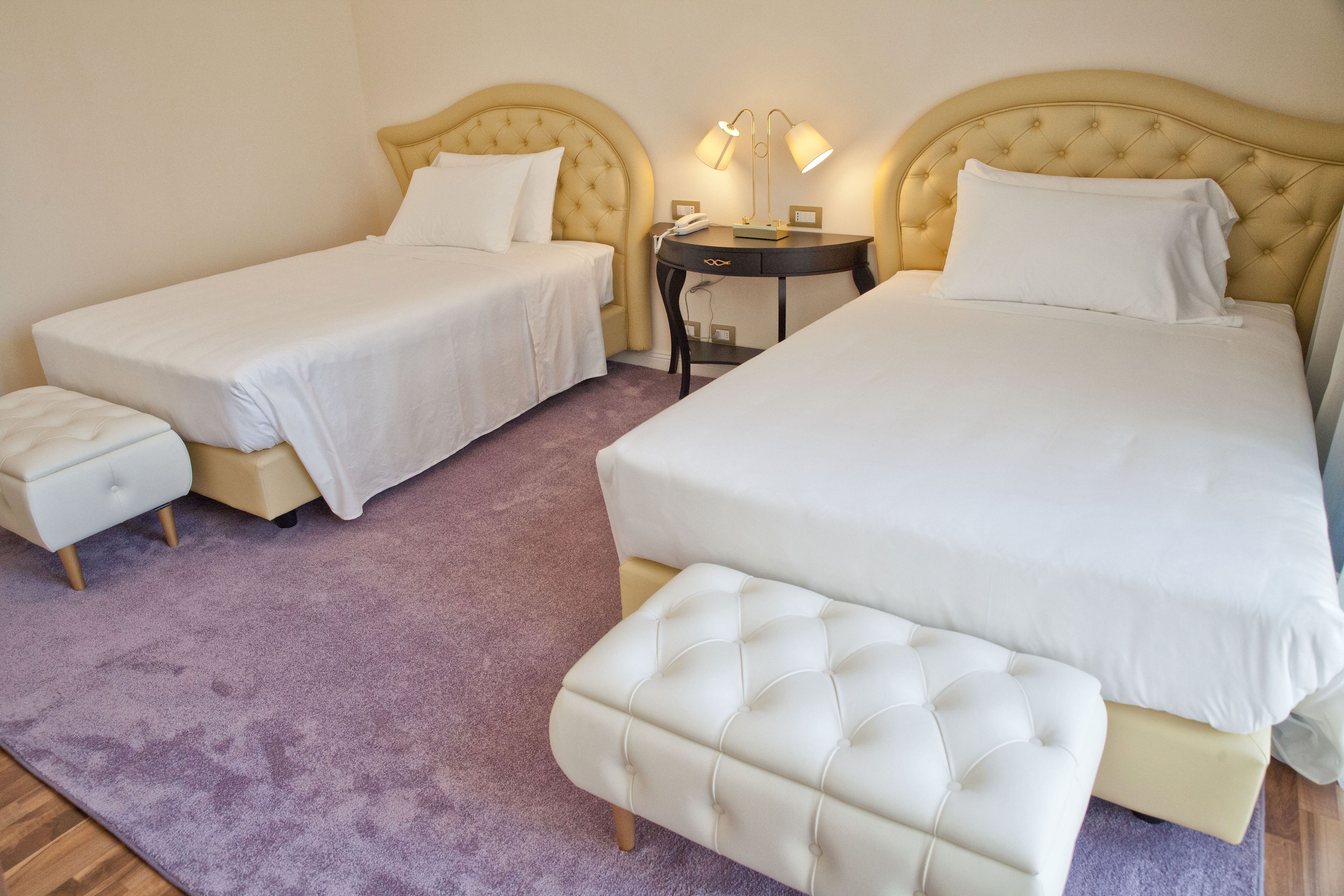 A hotel room with two single beds, a double bed, a small bedside table with lamps, and a matching ottoman.
