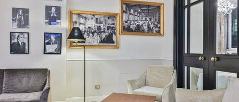 Una habitación con un sofá gris, un sillón beige, una mesa de centro de madera y una lámpara de pie negra. En la pared, fotografías enmarcadas en blanco y negro.