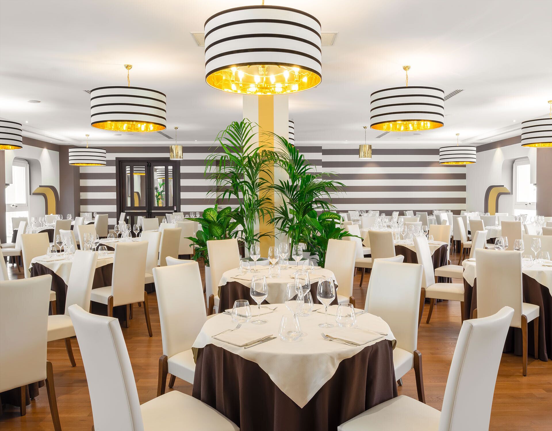 A restaurant with round tables set for dining, featuring striped ceiling lights and potted plants.