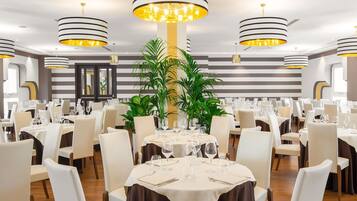 A restaurant with round tables set for dining, featuring striped ceiling lights and potted plants.