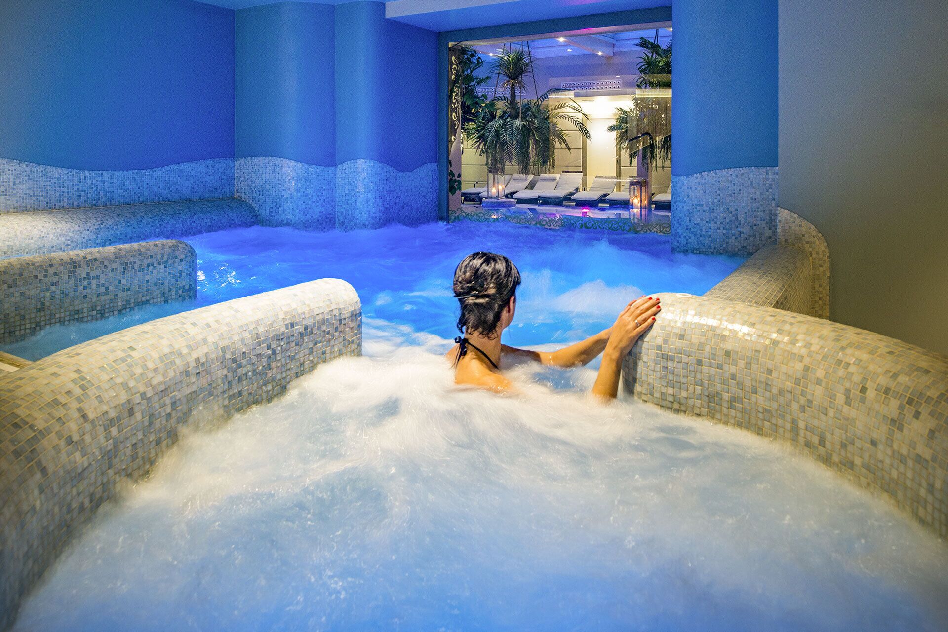A person relaxing in a hot tub with mosaic tiles, surrounded by blue-tinted walls and a view of palm trees.