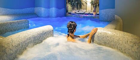 A person relaxing in a hot tub with mosaic tiles, surrounded by blue-tinted walls and a view of palm trees.