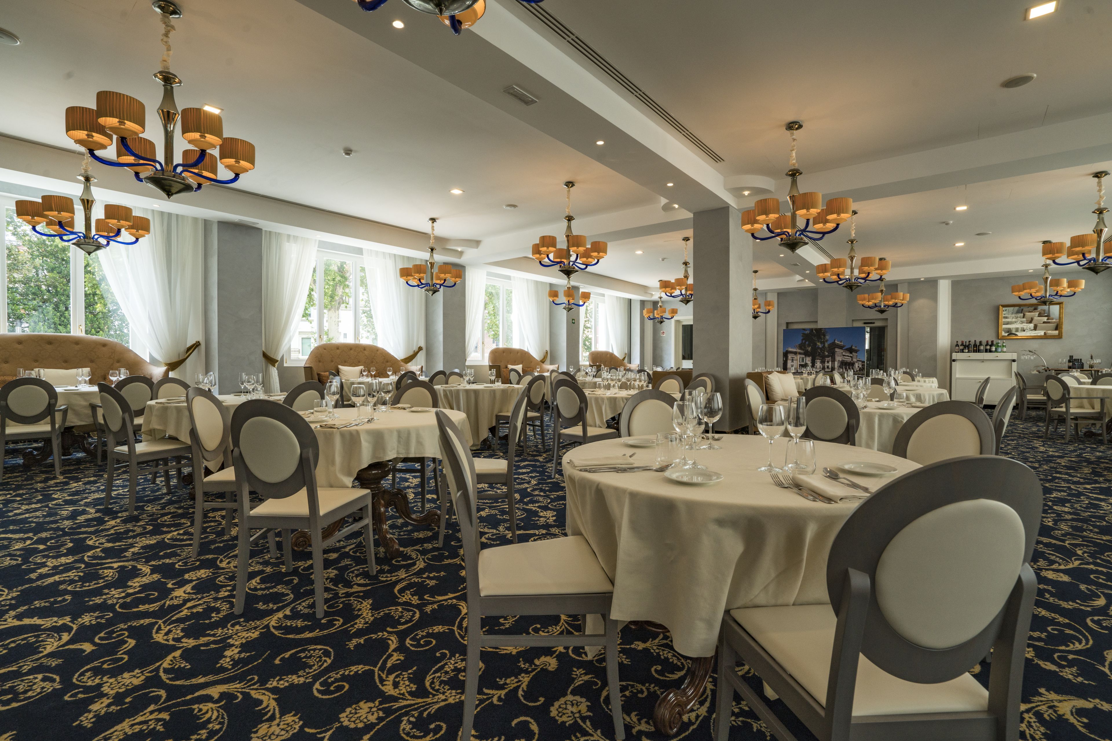 A formal dining room with multiple round tables, each set with white tablecloths and silverware. The room features ornate chandeliers and a patterned carpet.