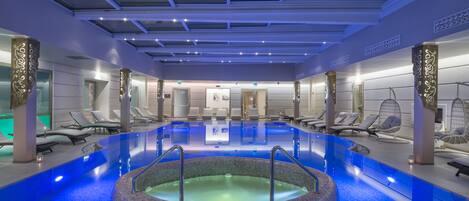 Una piscina interior moderna con un jacuzzi central, rodeada de sillones de descanso y un techo con tragaluces.
