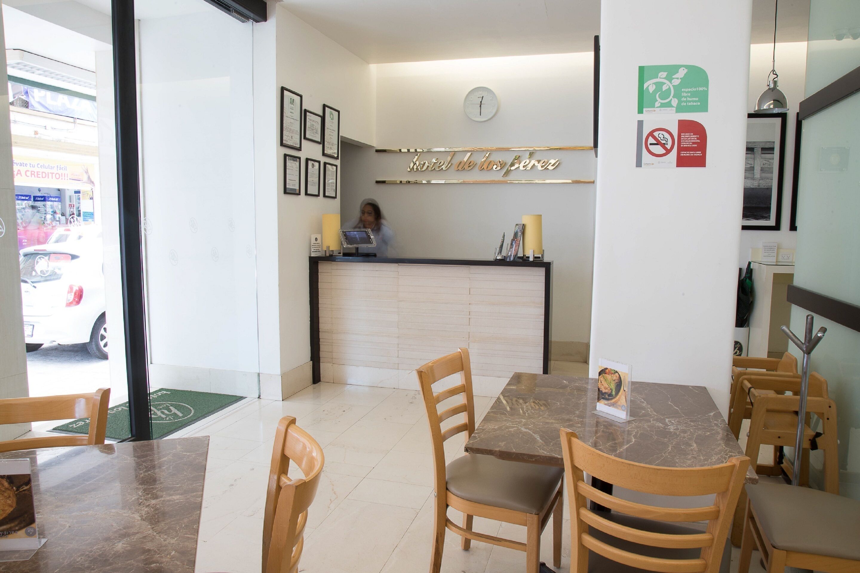 A hotel reception area with a reception desk, framed certificates, a clock, and a green mat at the entrance.