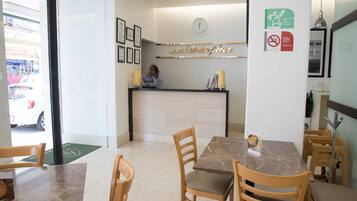 A hotel reception area with a reception desk, framed certificates, a clock, and a green mat at the entrance.