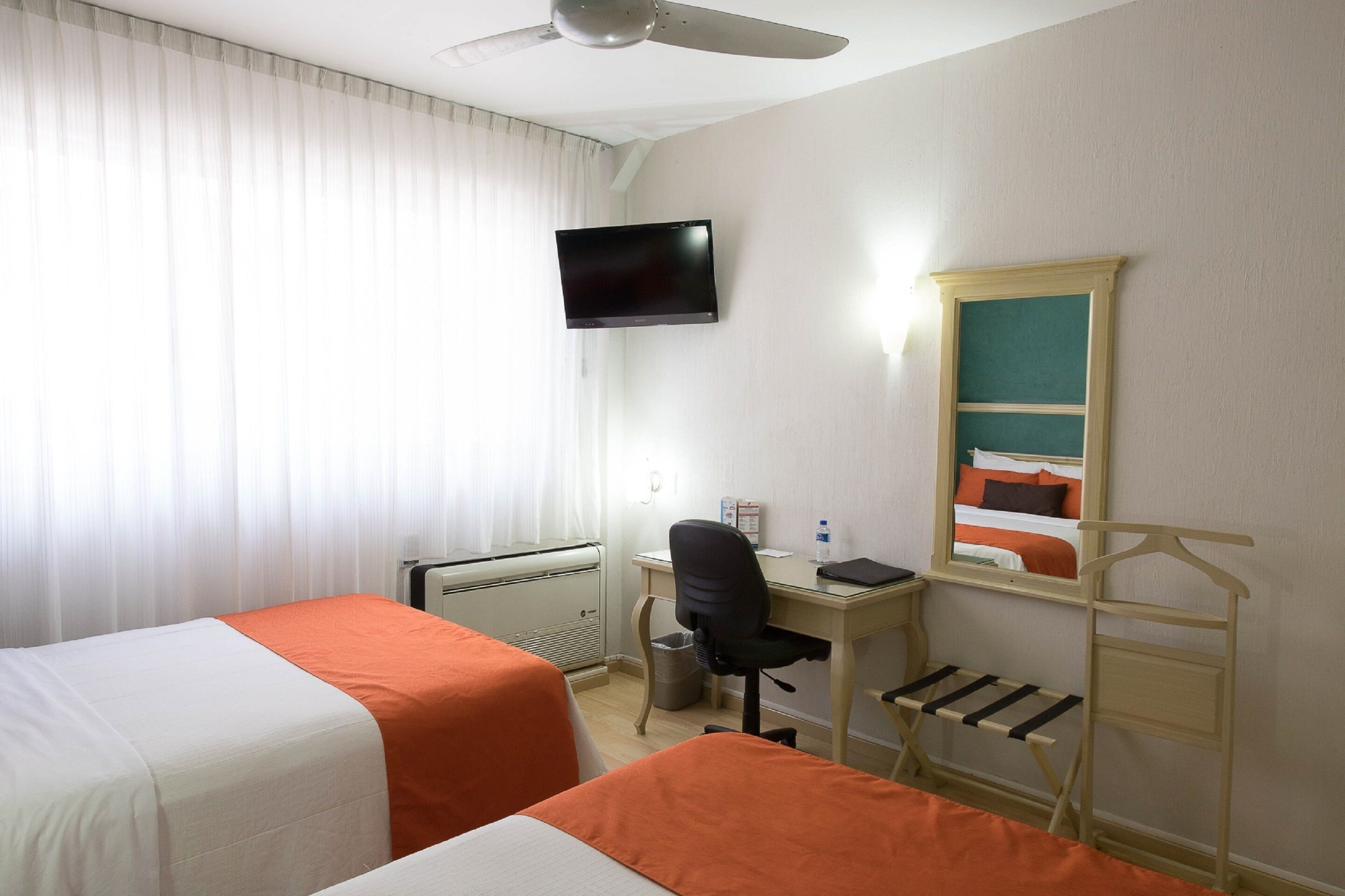 Ett hotellrum med två sängar, ett skrivbord med en stol, en TV, ett element och ett fönster med gardiner.