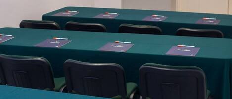Uma sala de conferências com mesas cobertas por toalhas verdes, cada uma com uma placa de identificação e um laptop. Uma tela de televisão exibe o logotipo do Piedra Alta Hotels.