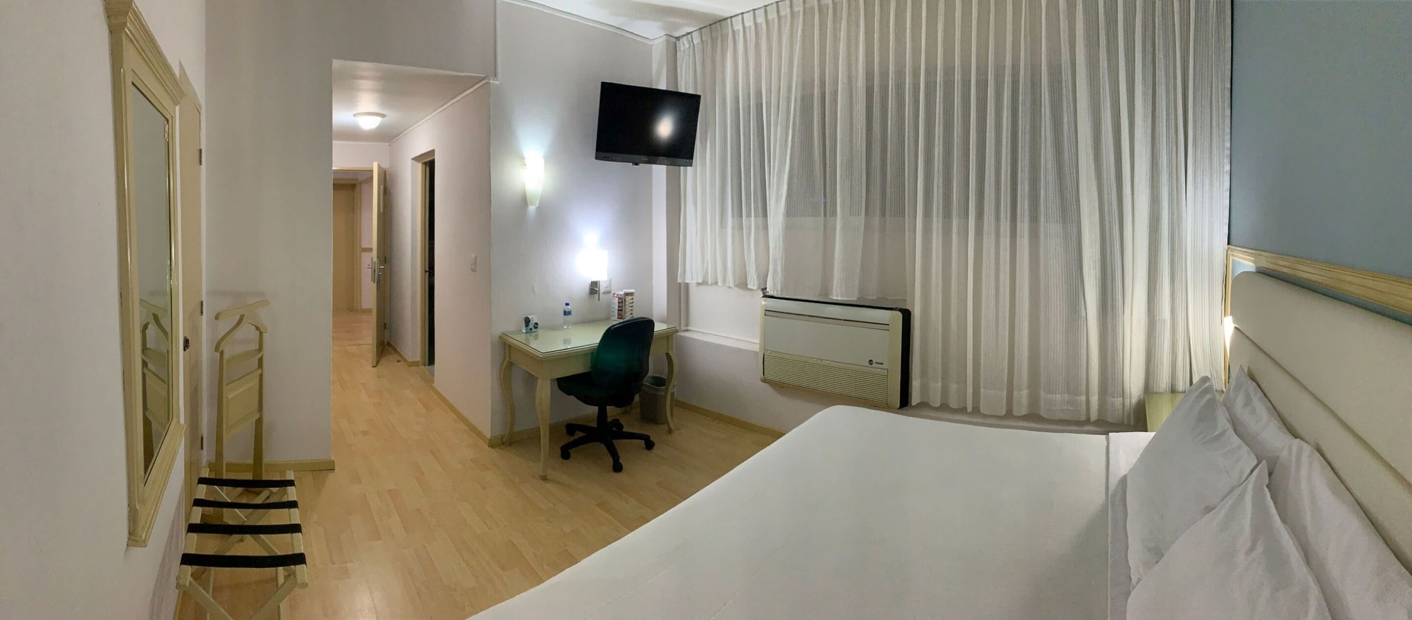 A hotel room with a bed, a desk with a chair, a television mounted on the wall, and a window with sheer curtains.