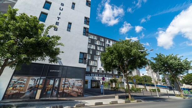 A modern hotel building with 'ALL SUITES' signage, a visible entrance, and a clear sky.