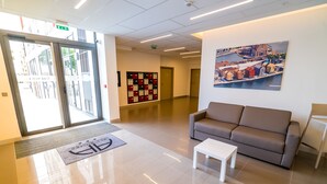 A lobby with a glass door entrance, a beige sofa, a white coffee table, a large framed cityscape painting, and a row of lockers.