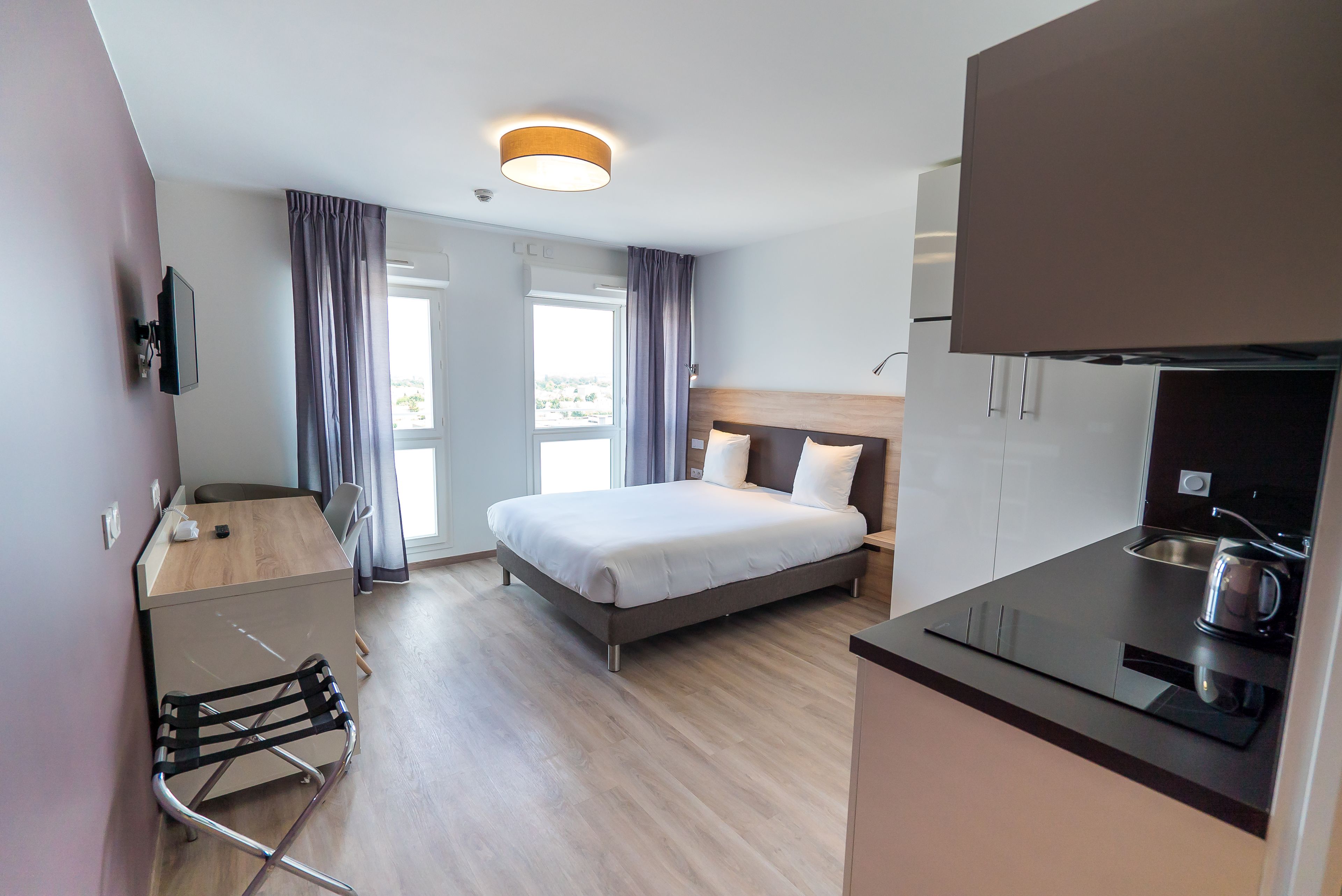 A modern hotel room with a bed, a desk, a chair, a kitchenette, and a view of the outdoors.