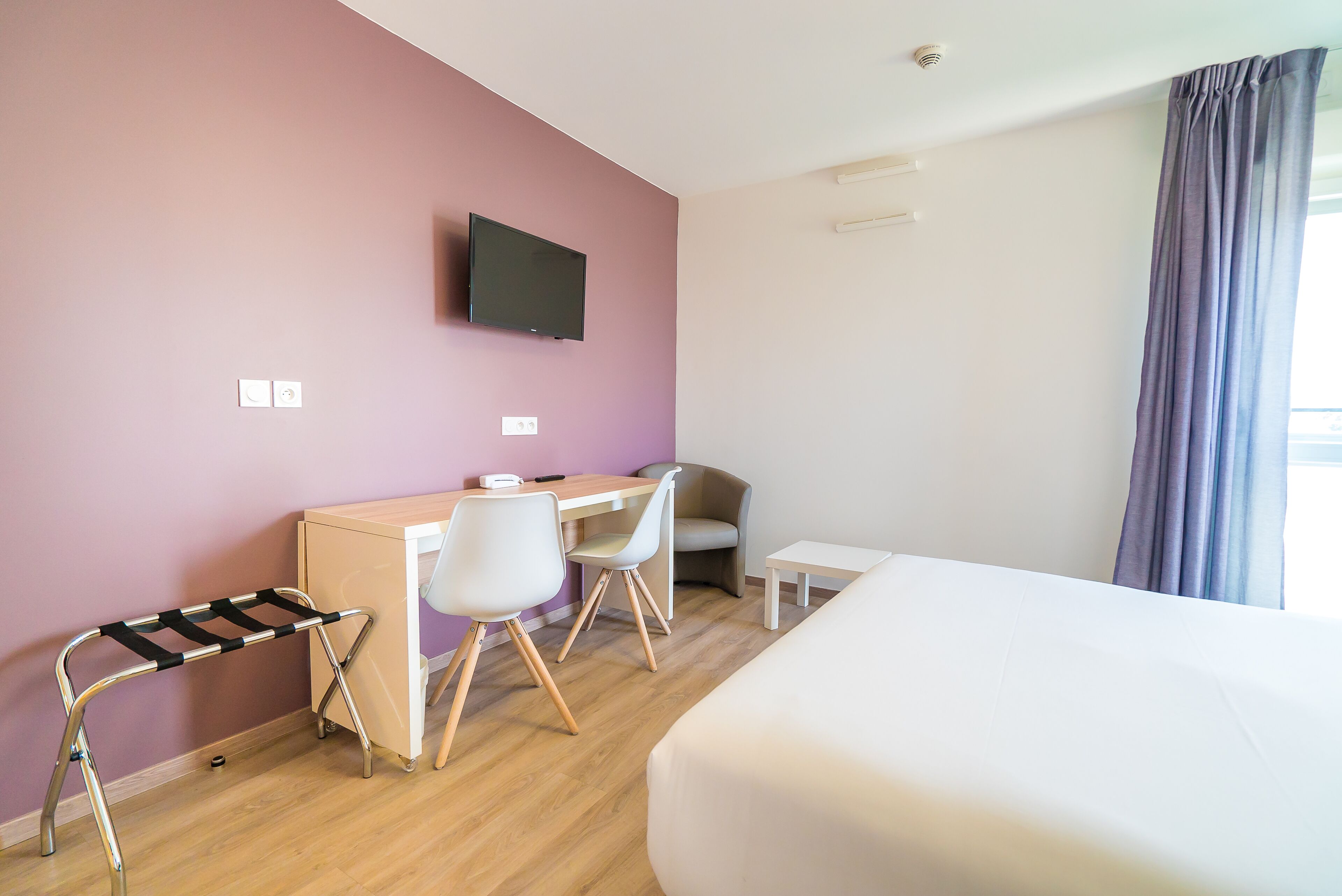 Une chambre d’hôtel comprenant un lit, un bureau avec deux chaises, un téléviseur fixé au mur et une fenêtre avec des rideaux.