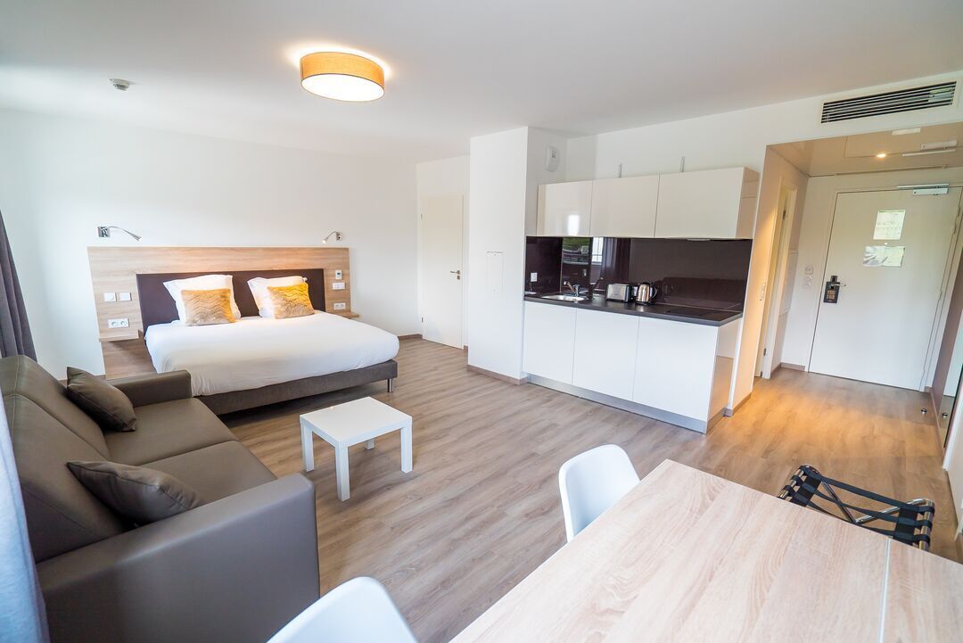 A modern hotel room with a bed, sofa, dining table, and kitchenette.