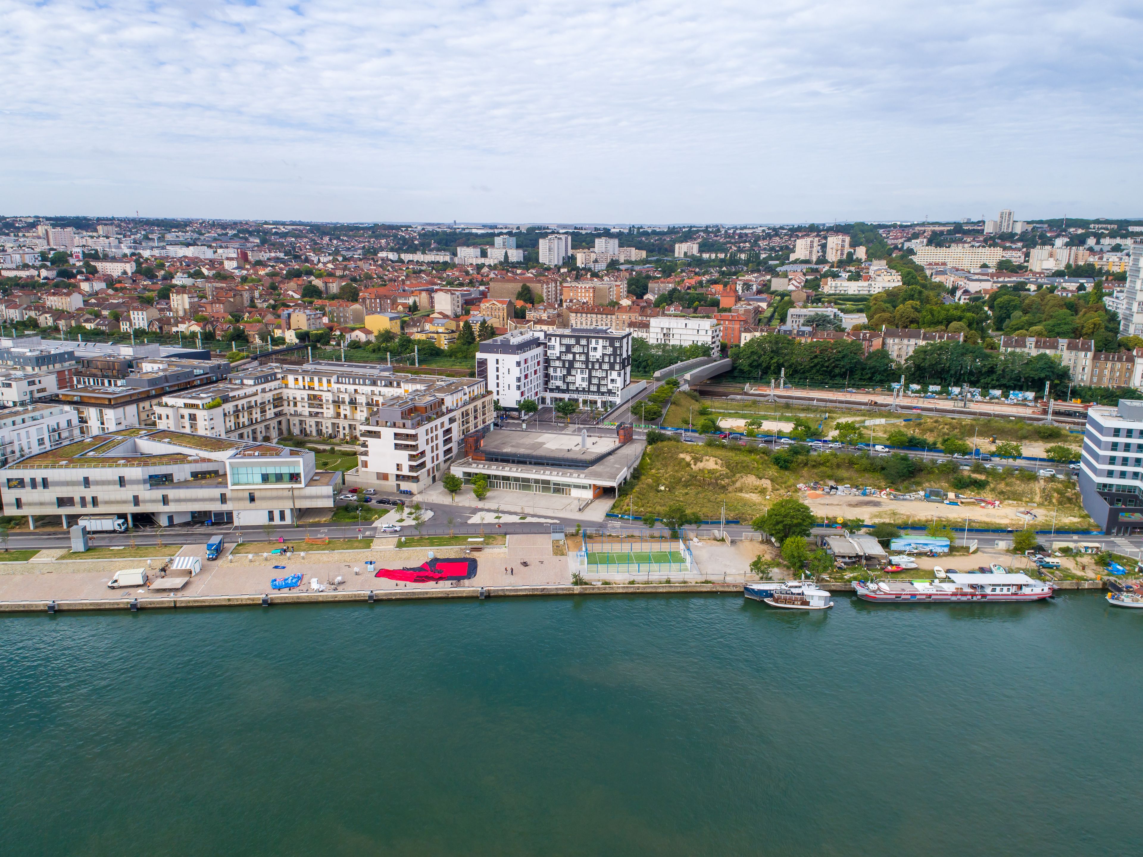 Stadsgezicht aan het water met moderne gebouwen, een jachthaven met boten en een voetpad langs de rivier.