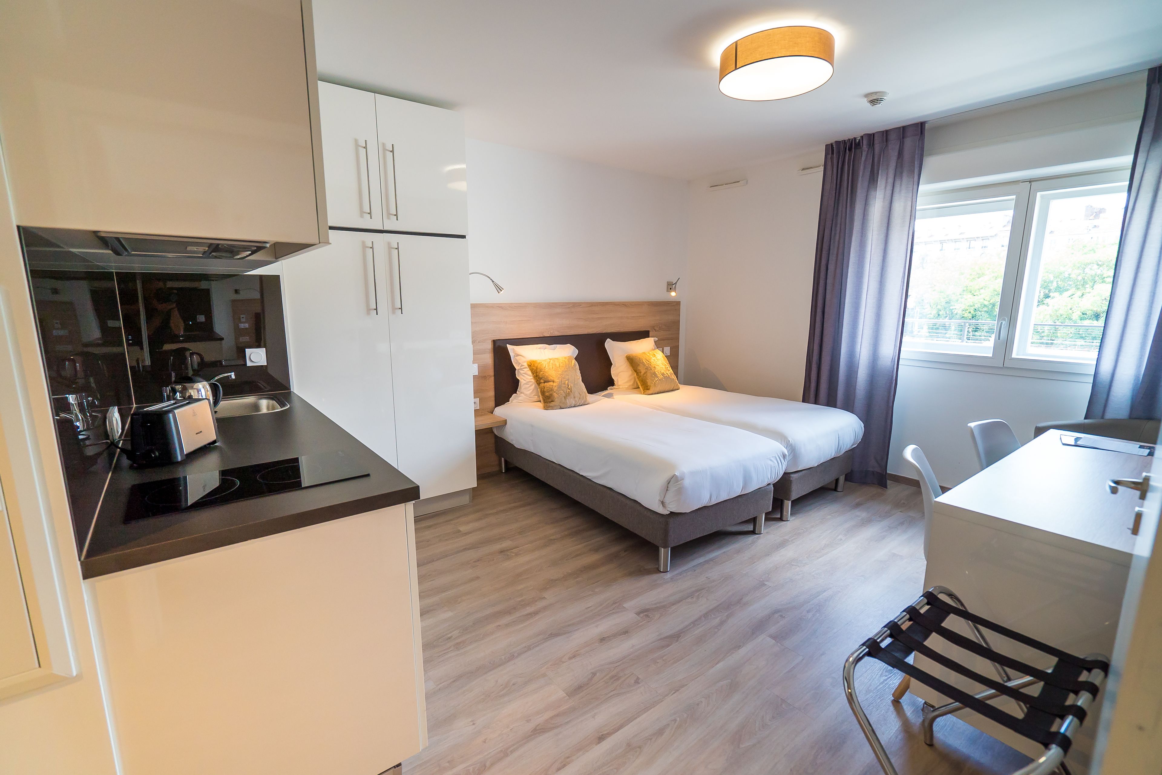 Una habitación de hotel moderna con kitchenette, una cama con dos almohadas, un escritorio y una ventana con cortinas.
