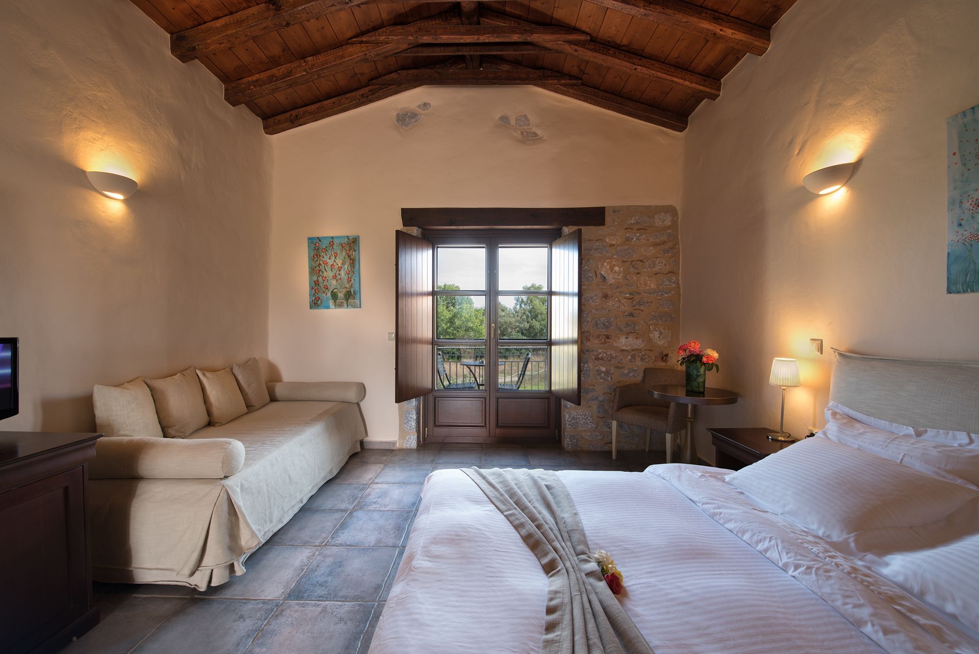 A bedroom with a large bed, a sofa, a small table with a lamp, and a view through a door.