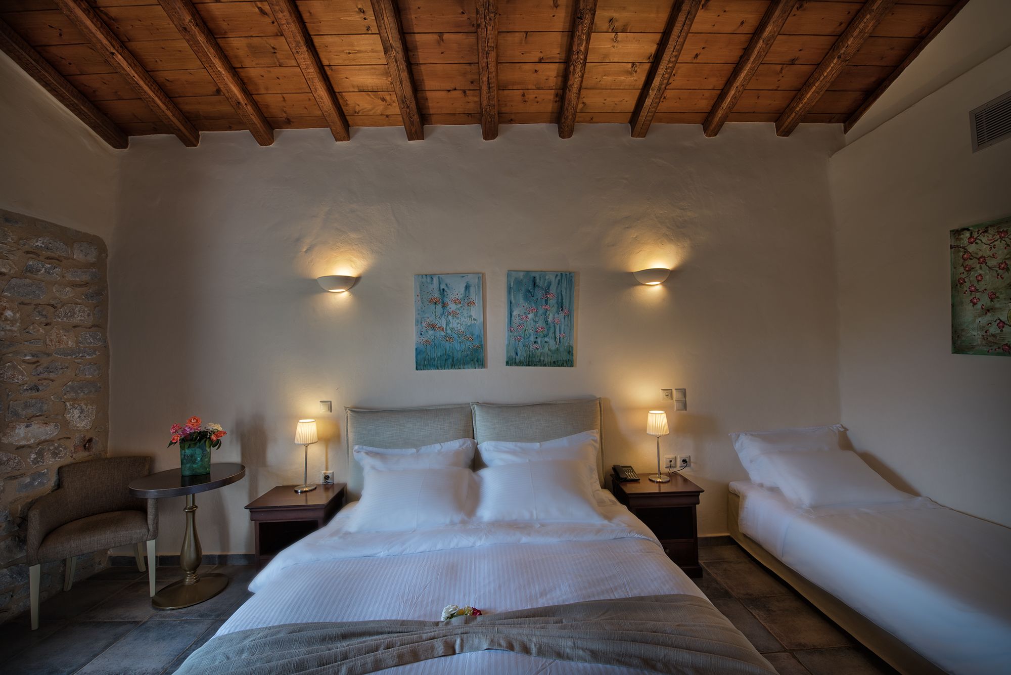 A bedroom with a large bed, two bedside tables, a chair, and a stone wall.
