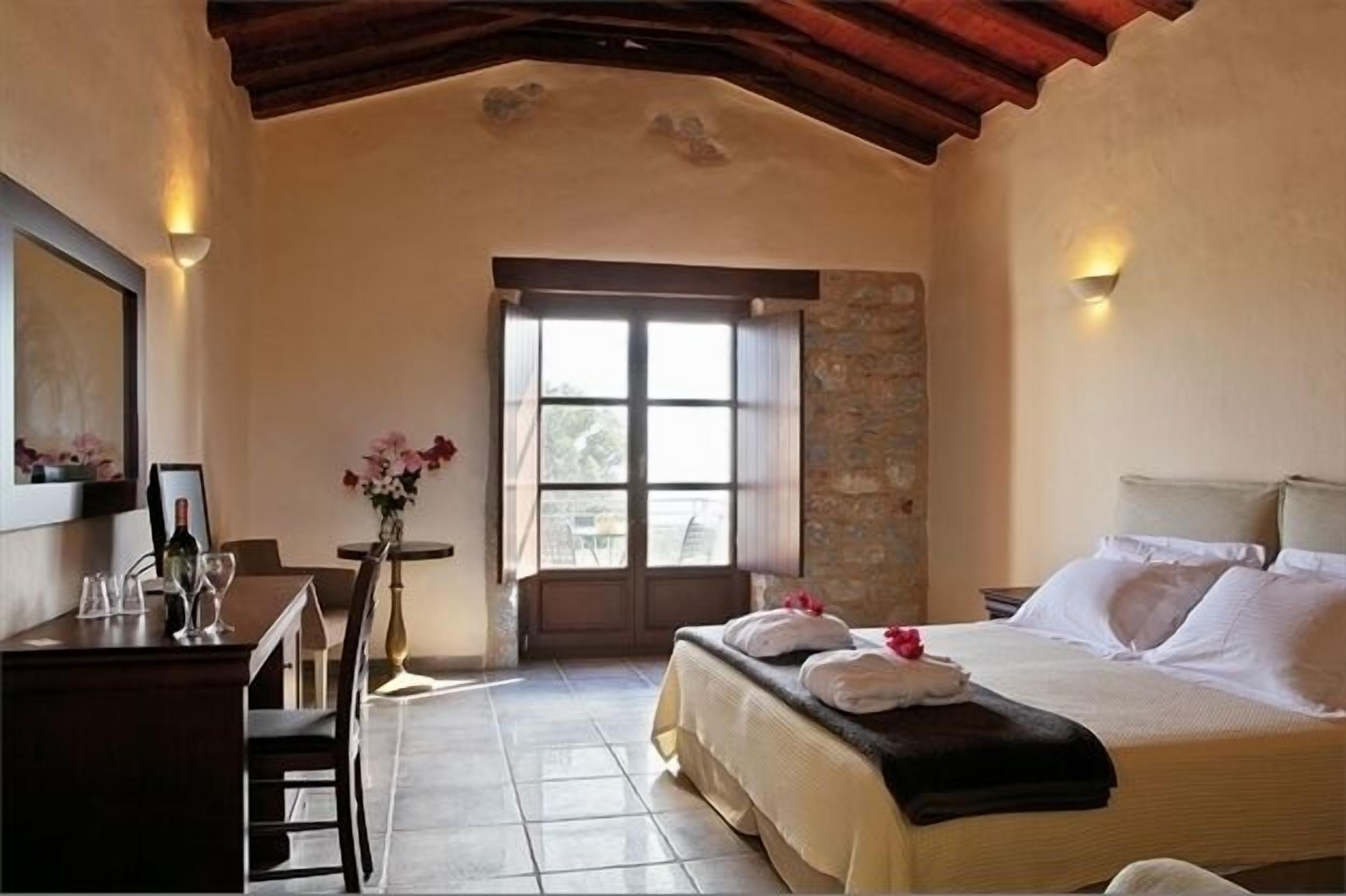 A bedroom with a bed, a desk with a chair, a window, and a stone wall.