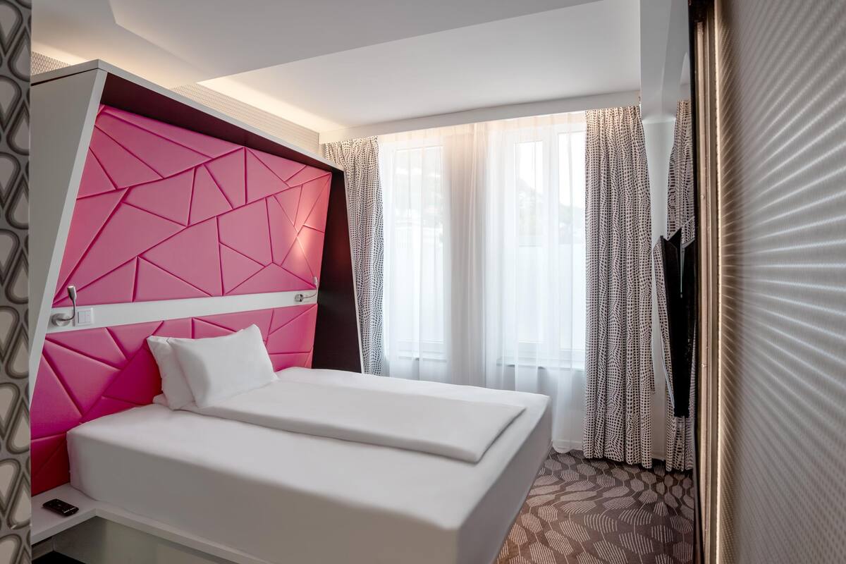 A hotel room with a bed, a pink accent wall, a window with curtains, and a patterned carpet.