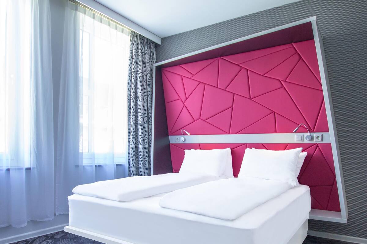 A modern hotel room with a large bed, a geometric patterned headboard, and a view of the outdoors through sheer curtains.