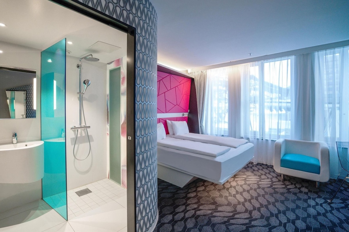 A modern hotel room with a glass-enclosed shower, a bed with a pink headboard, a white sofa, and a view of the city through large windows.