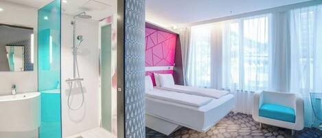 A modern hotel room with a glass-enclosed shower, a large bed with a geometric headboard, a white sofa, and a view of the outdoors.
