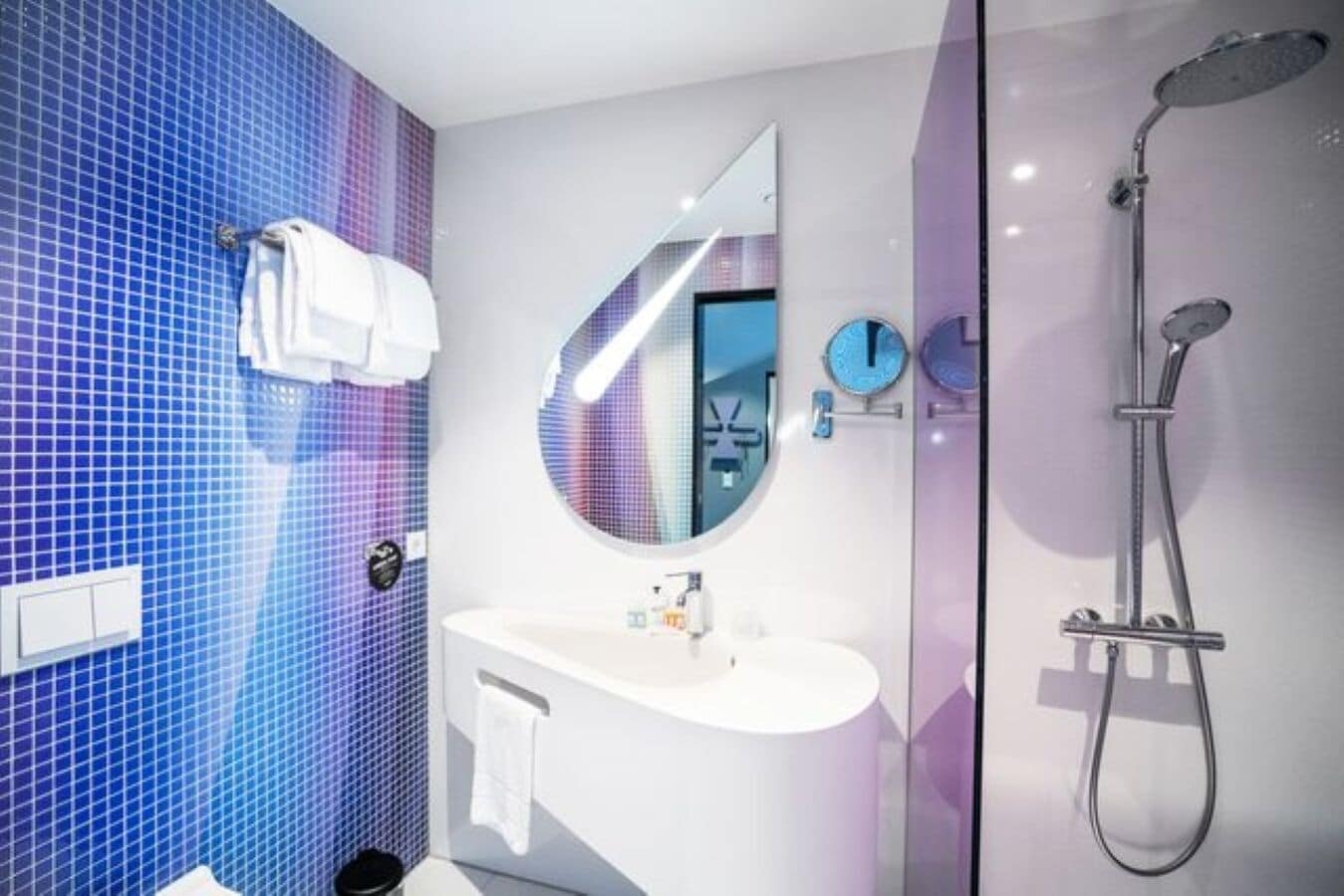 Un baño moderno con pared de azulejos azules, un espejo redondo, un lavabo blanco y una ducha con mampara de vidrio.