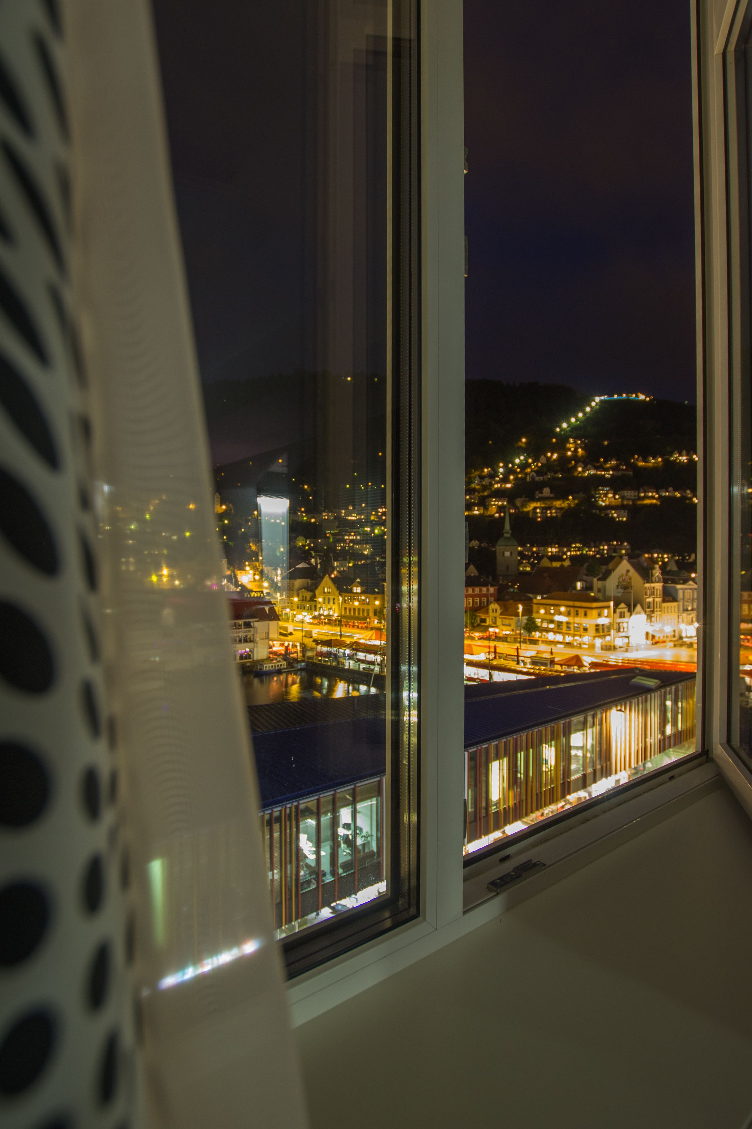 Une vue depuis une fenêtre donnant sur un paysage urbain nocturne, avec des bâtiments illuminés et une colline lointaine.